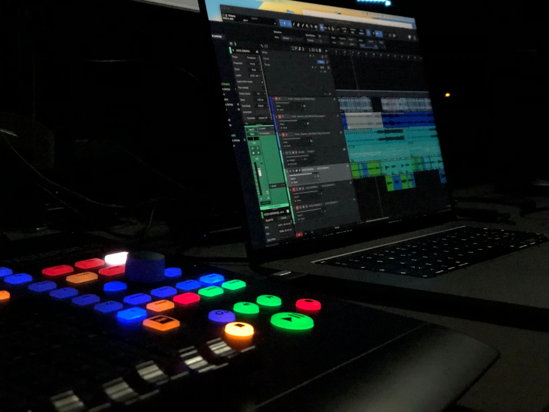 Dark studio setup with a laptop showing music editing software and a control pad with colorful buttons.
